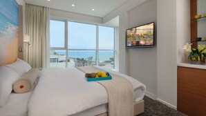 Habitación de hotel con una cama grande, un televisor montado en la pared, un ventanal con vista al océano y una mesita con una planta y una bandeja de fruta.