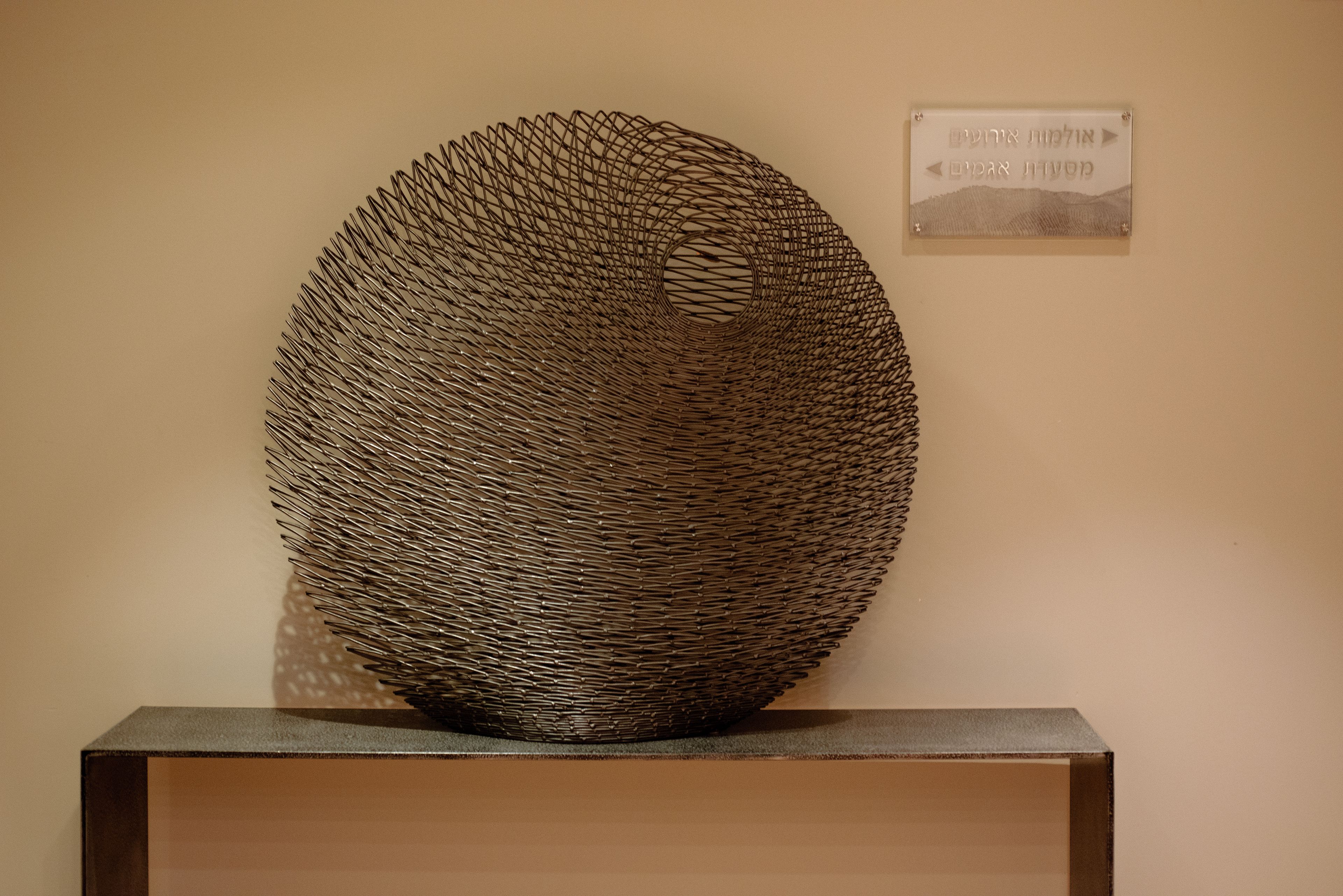 Una escultura metálica esférica sobre un pedestal frente a una pared color beige.