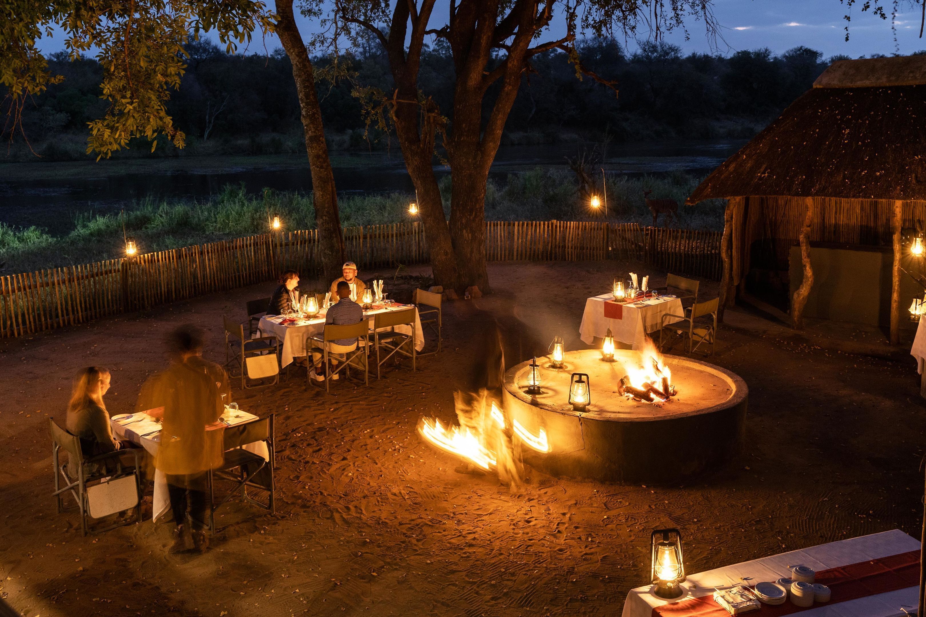 An outdoor dining area with tables and chairs, a central fire pit, and a rustic thatched-roof structure.