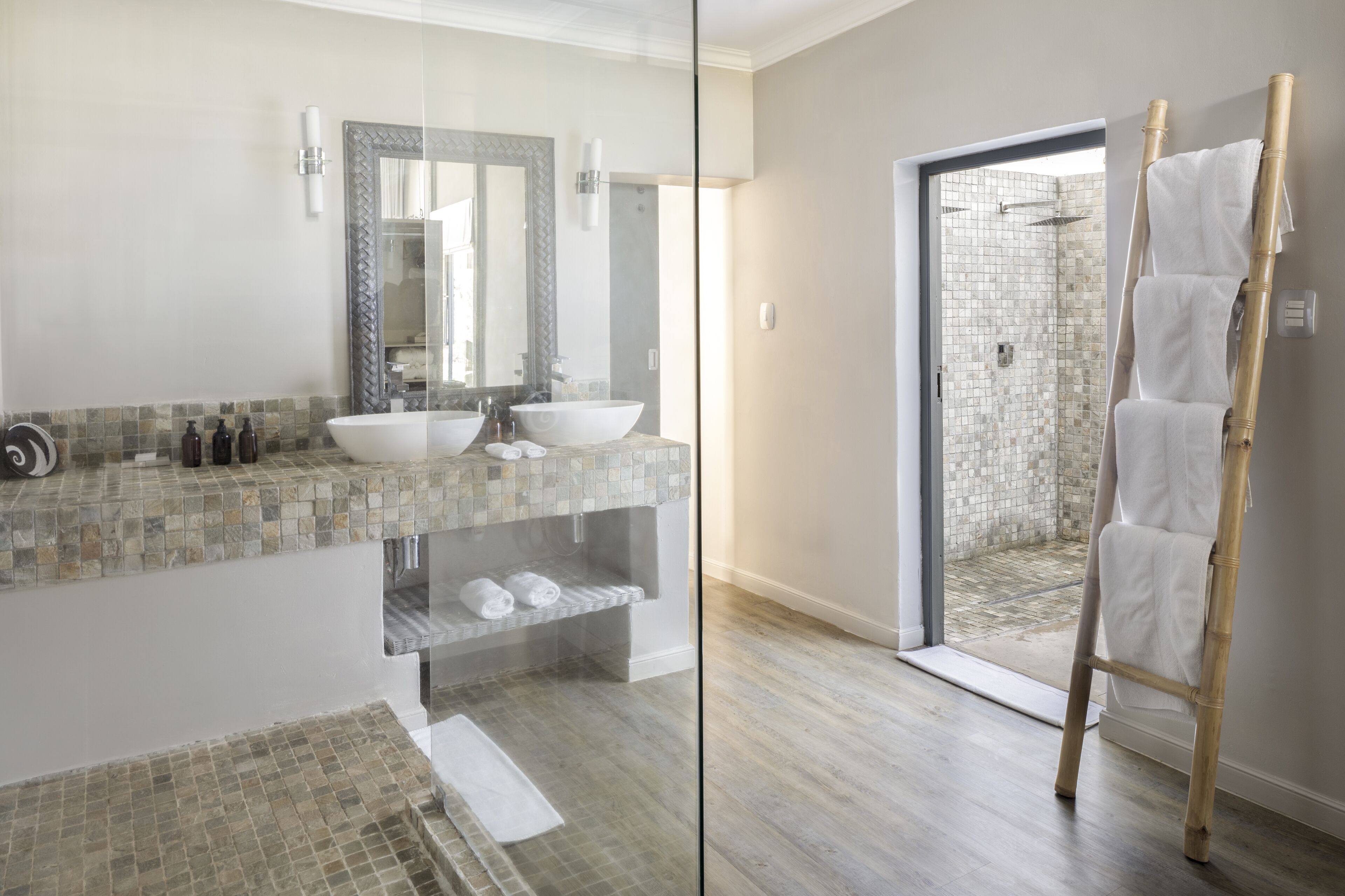 Een badkamer met een glazen scheidingswand, een grote spiegel, twee wasbakken, een douchegedeelte en een bamboe handdoekhouder.