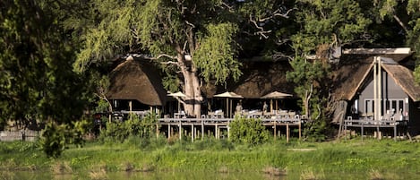 A riverside dining area with thatched-roof tables and umbrellas, surrounded by lush greenery and trees.