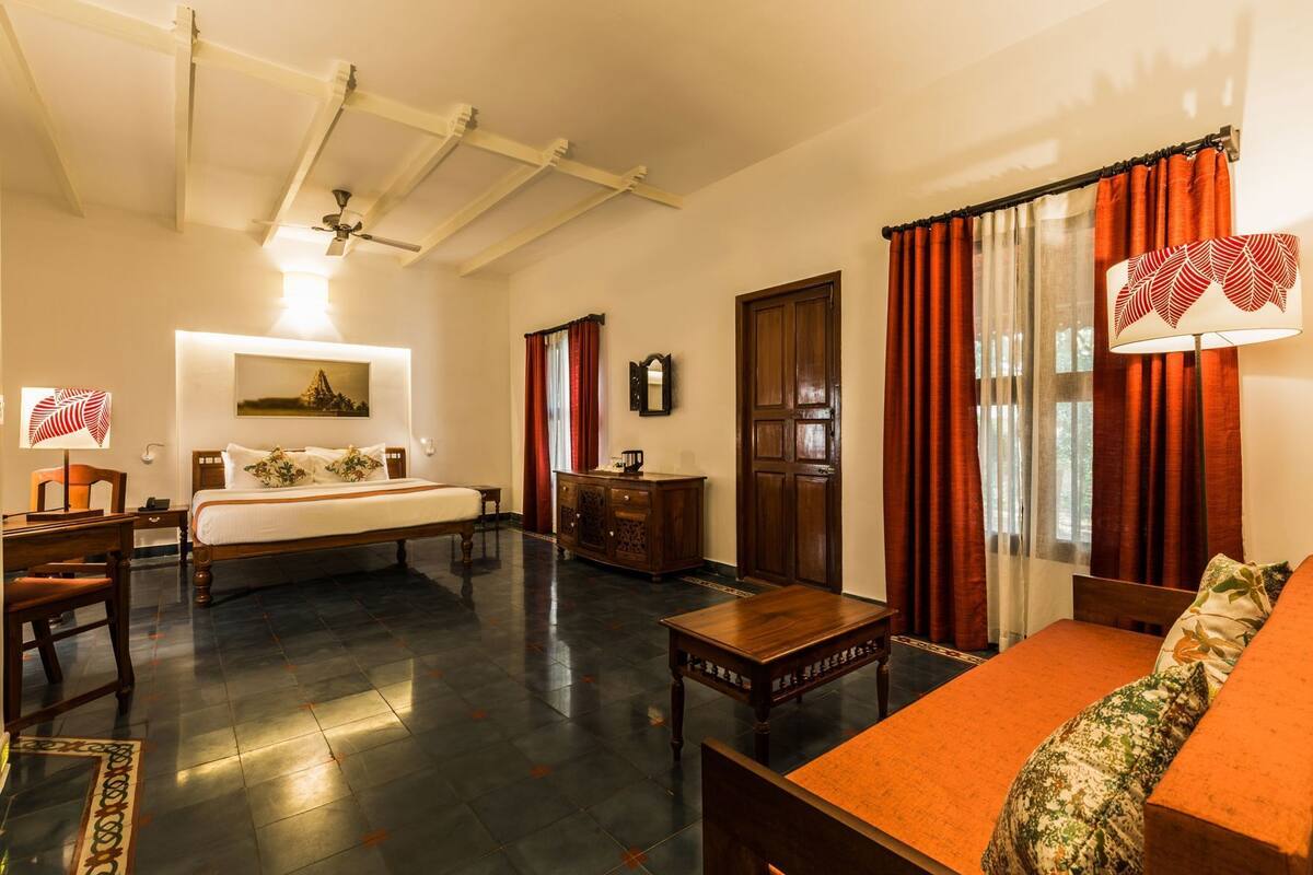 Une chambre d’hôtel comprenant un lit, un bureau, un canapé, une armoire en bois, un miroir et un ventilateur de plafond.
