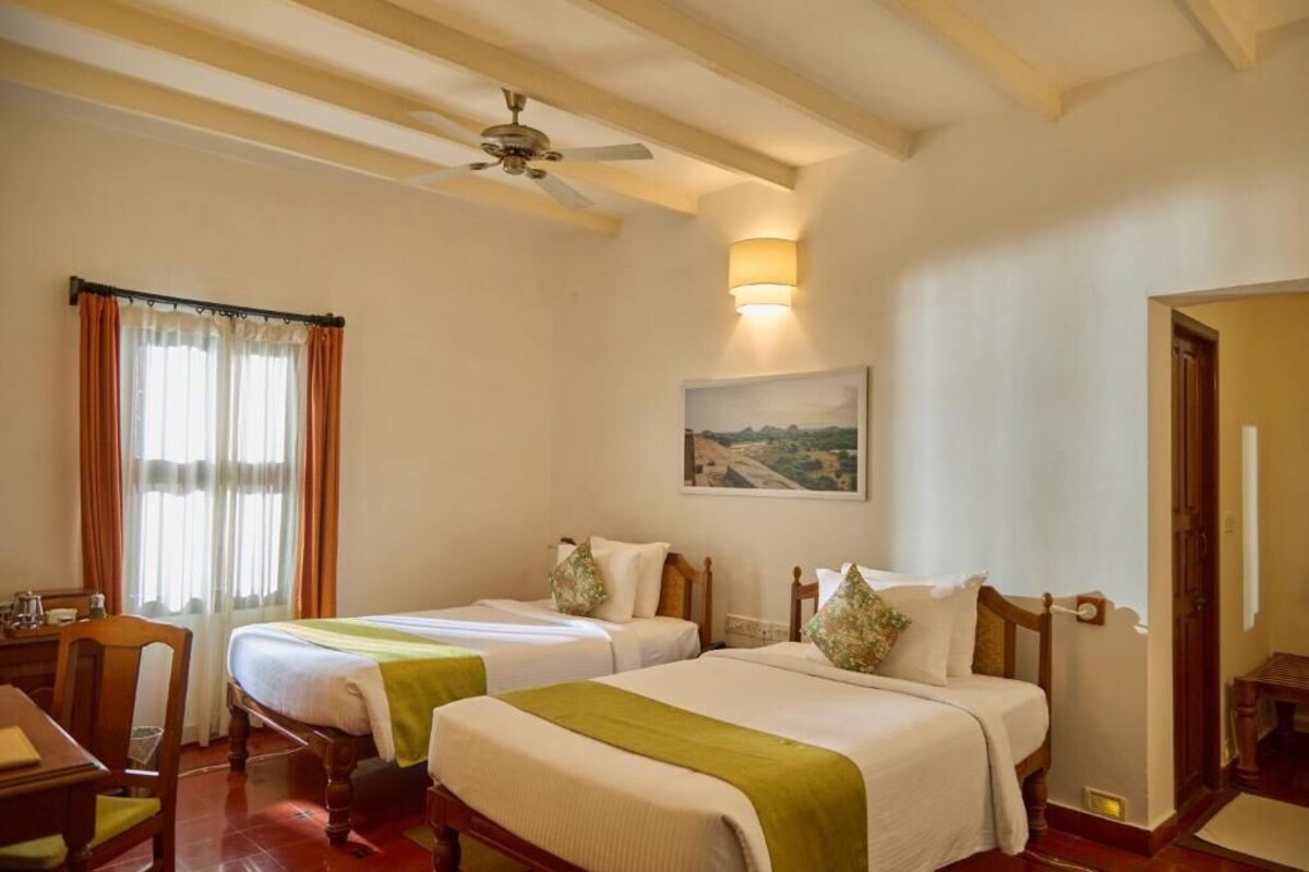 A hotel room with two beds, a ceiling fan, a painting on the wall, and a window with curtains.