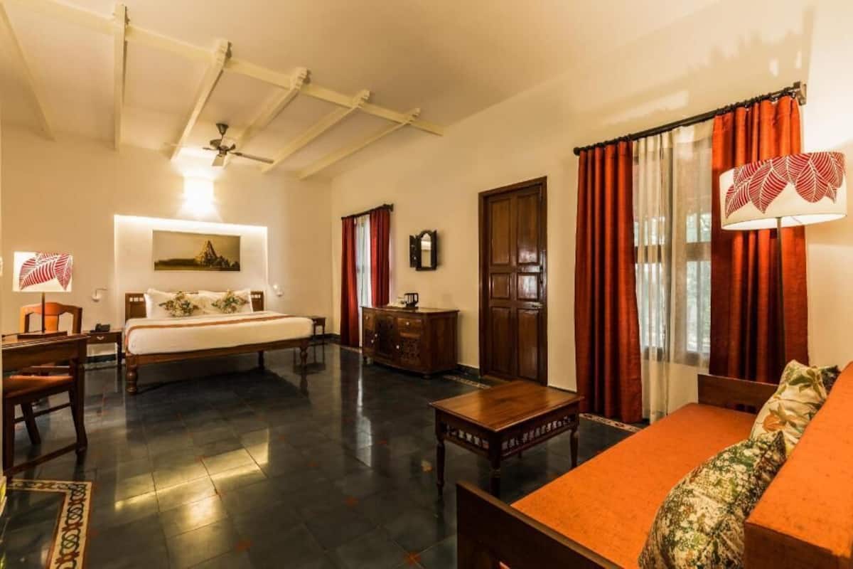Une chambre d’hôtel comprenant un lit, un canapé, une table en bois, un bureau, une chaise, un ventilateur de plafond, une horloge murale et une fenêtre avec des rideaux.