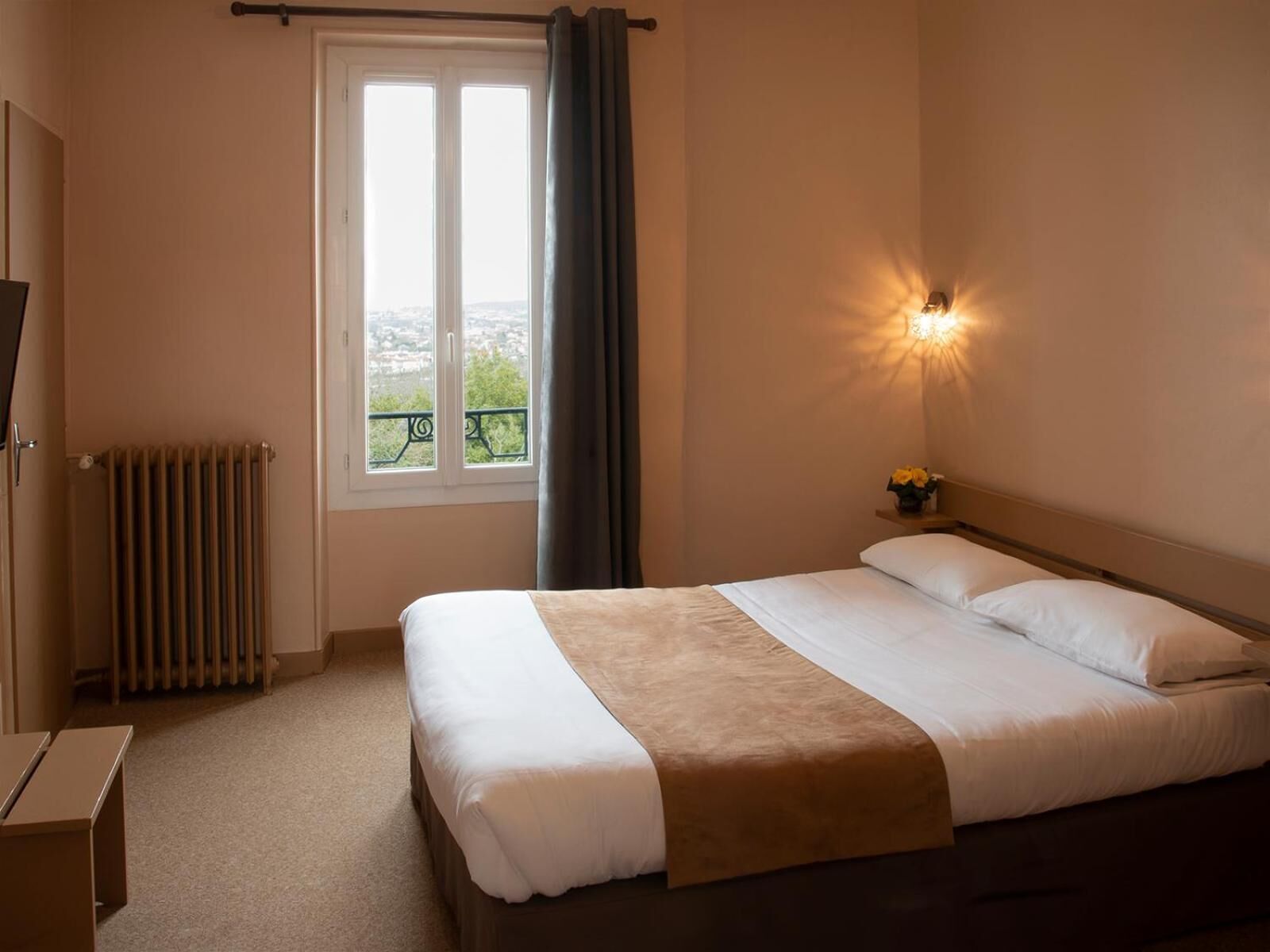 Een hotelkamer met een bed, nachtkastje met lamp, raam met gordijnen en een radiator.