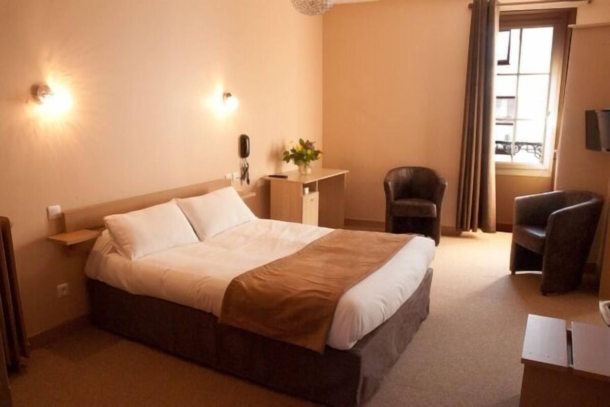 Une chambre d’hôtel avec un grand lit, un bureau avec un téléphone, une chaise et une fenêtre avec des rideaux.