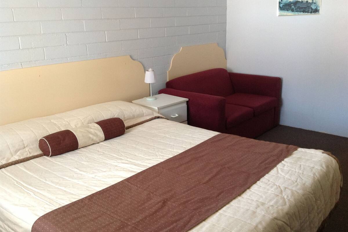 A hotel room with a bed, a nightstand, a red chair, and a wall-mounted painting.