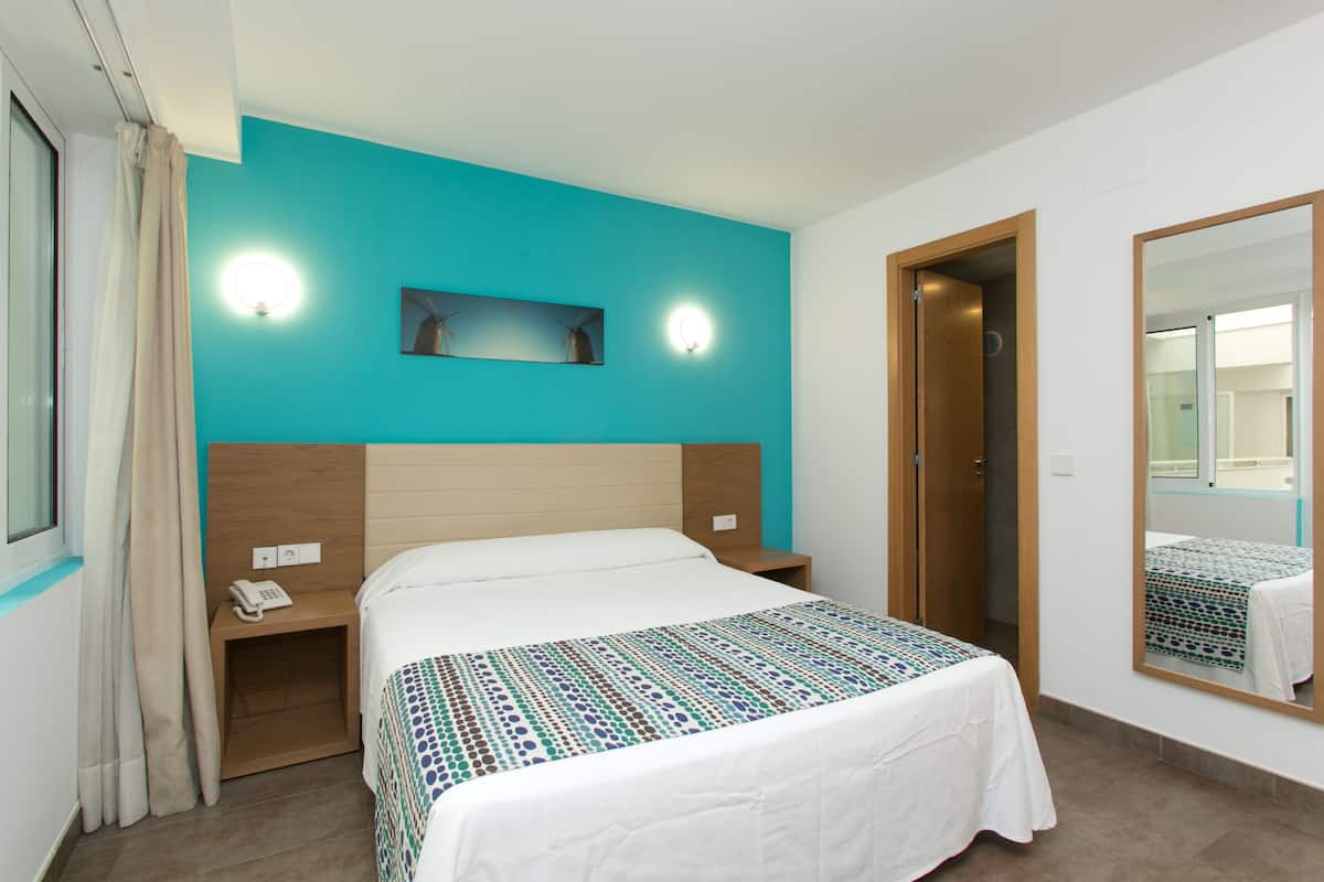 Quarto de hotel com cama, mesa de cabeceira, telefone, espelho e porta.