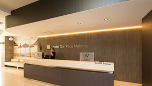 Un espace d’accueil d’hôtel au design moderne, comprenant un comptoir de réception, une enseigne indiquant « hôtel Don Pepe Majorque » et un coin salon offrant une vue sur la plage.