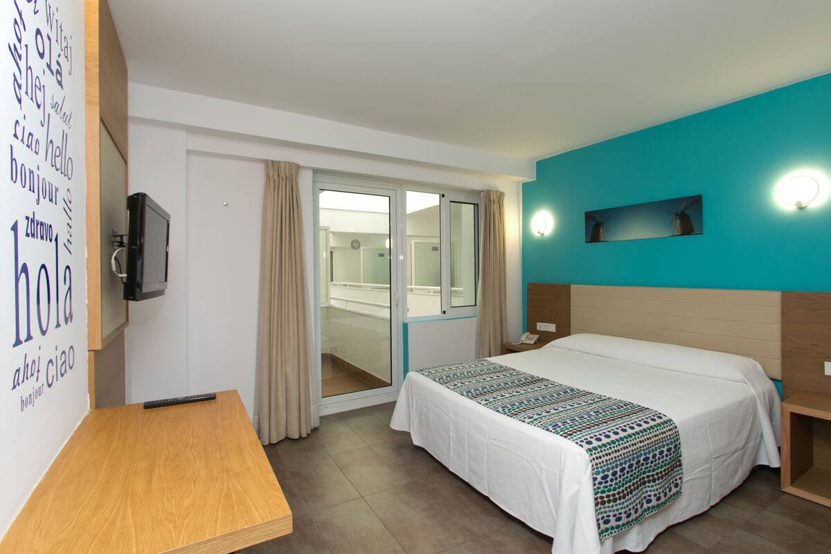 Quarto de hotel com cama, escrivaninha, televisão e porta de correr para uma varanda.