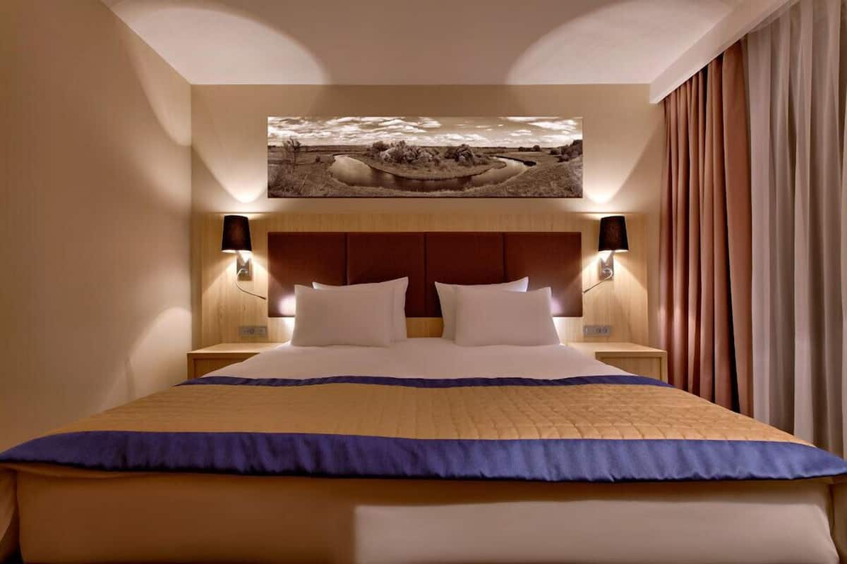Ein Hotelzimmer mit einem großen Bett, zwei Nachttischen mit Lampen, einem Vorhang mit Landschaftsbild und Blick ins Freie durch das Fenster.
