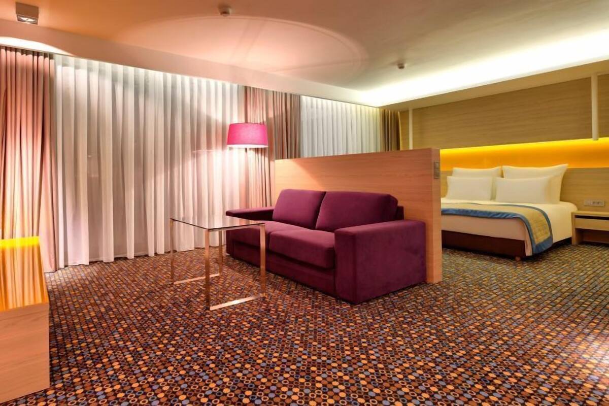Ein Hotelzimmer mit violettem Sofa, einem Bett mit weißer Bettwäsche, einem Holz-Kopfteil und einem gemusterten Teppich.