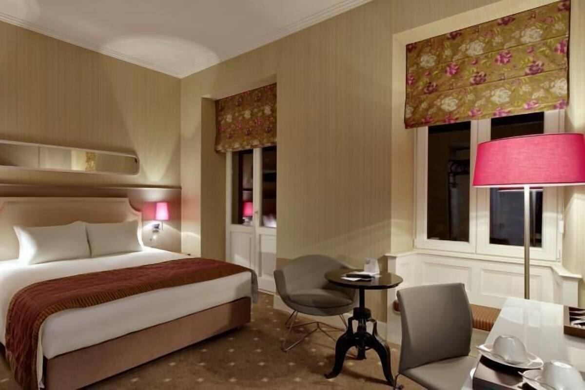 Ein Hotelzimmer mit einem großen Bett, zwei Nachttischlampen, einem kleinen Tisch mit Stuhl und einem Fenster mit Blumenmuster-Vorhang.