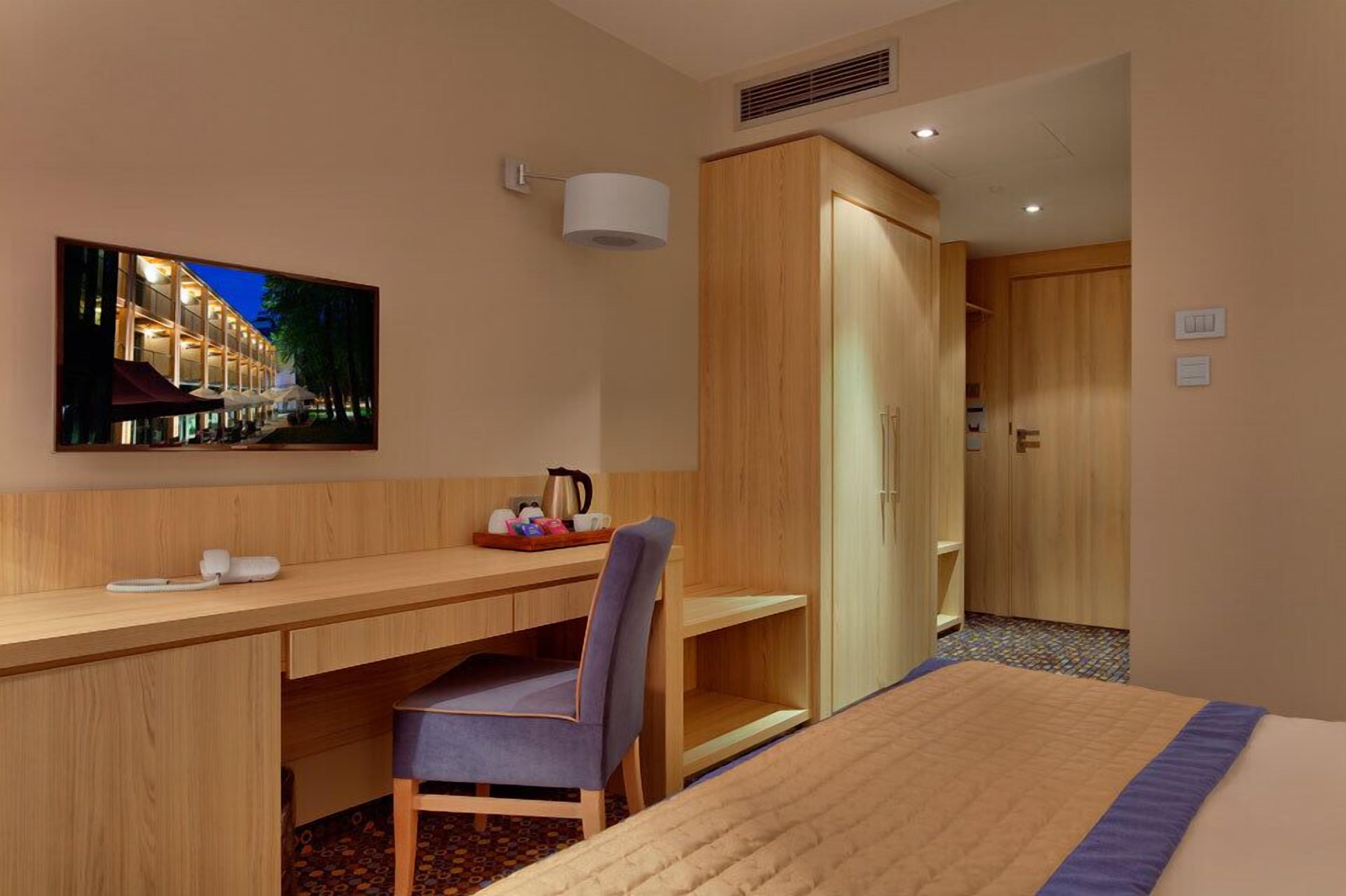 A hotel room with a wooden desk, a chair, a bed, a wardrobe, a television, and a coffee maker.