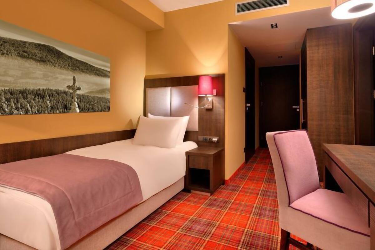 Ein Hotelzimmer mit Bett, Schreibtisch, Stuhl, Nachttisch, Lampe und einem Bild an der Wand.