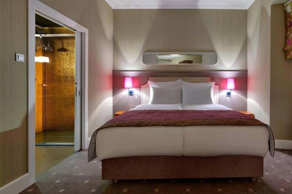 Ein Hotelzimmer mit einem großen Bett, zwei Nachttischlampen, einem Kopfteil mit Spiegel und einer Tür zu einem Badezimmer.