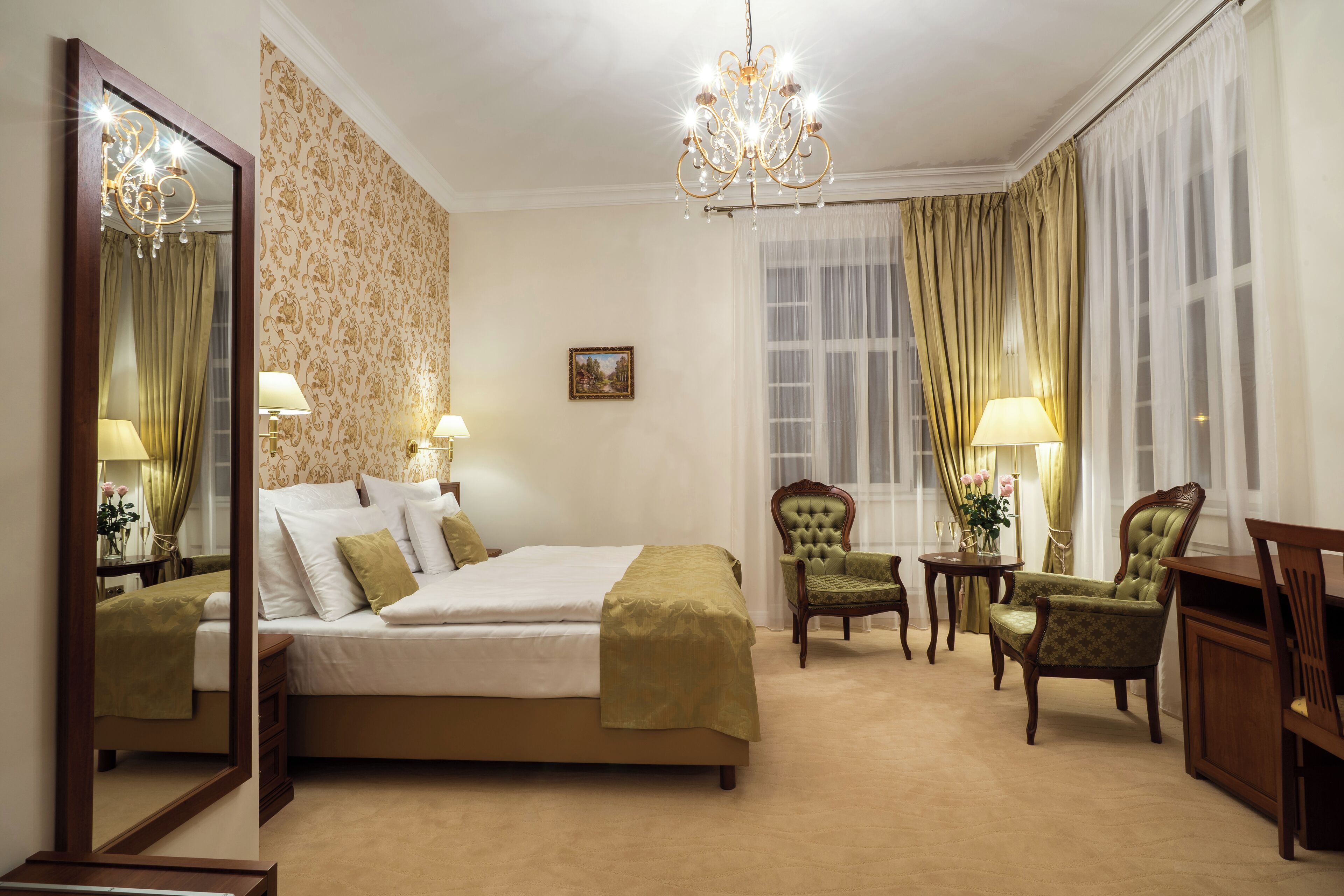 Habitación de hotel con una cama grande, dos sillones, un escritorio, una araña y un espejo amplio.