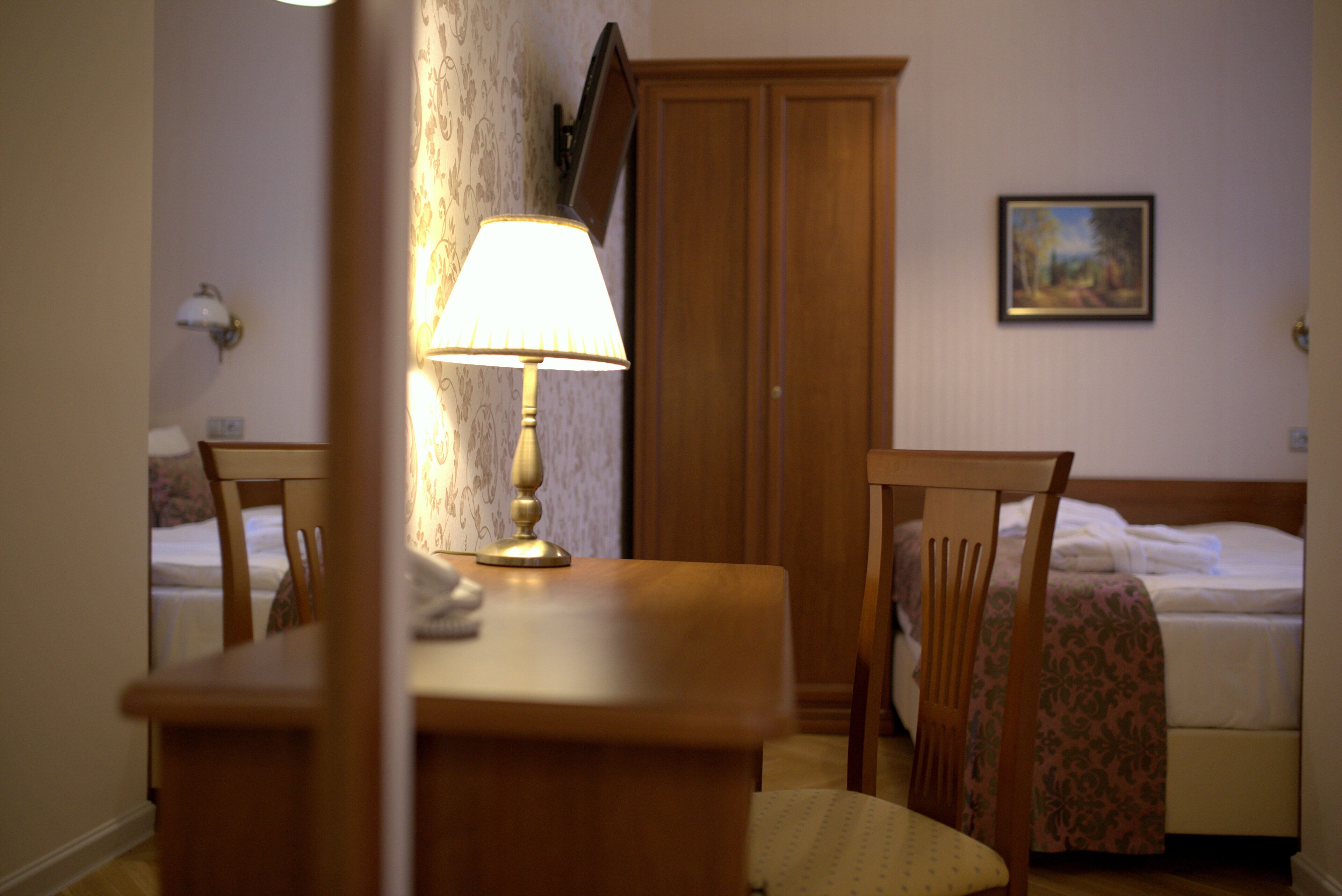 A hotel room with a wooden desk, a lamp, a chair, a bed, and a wardrobe.