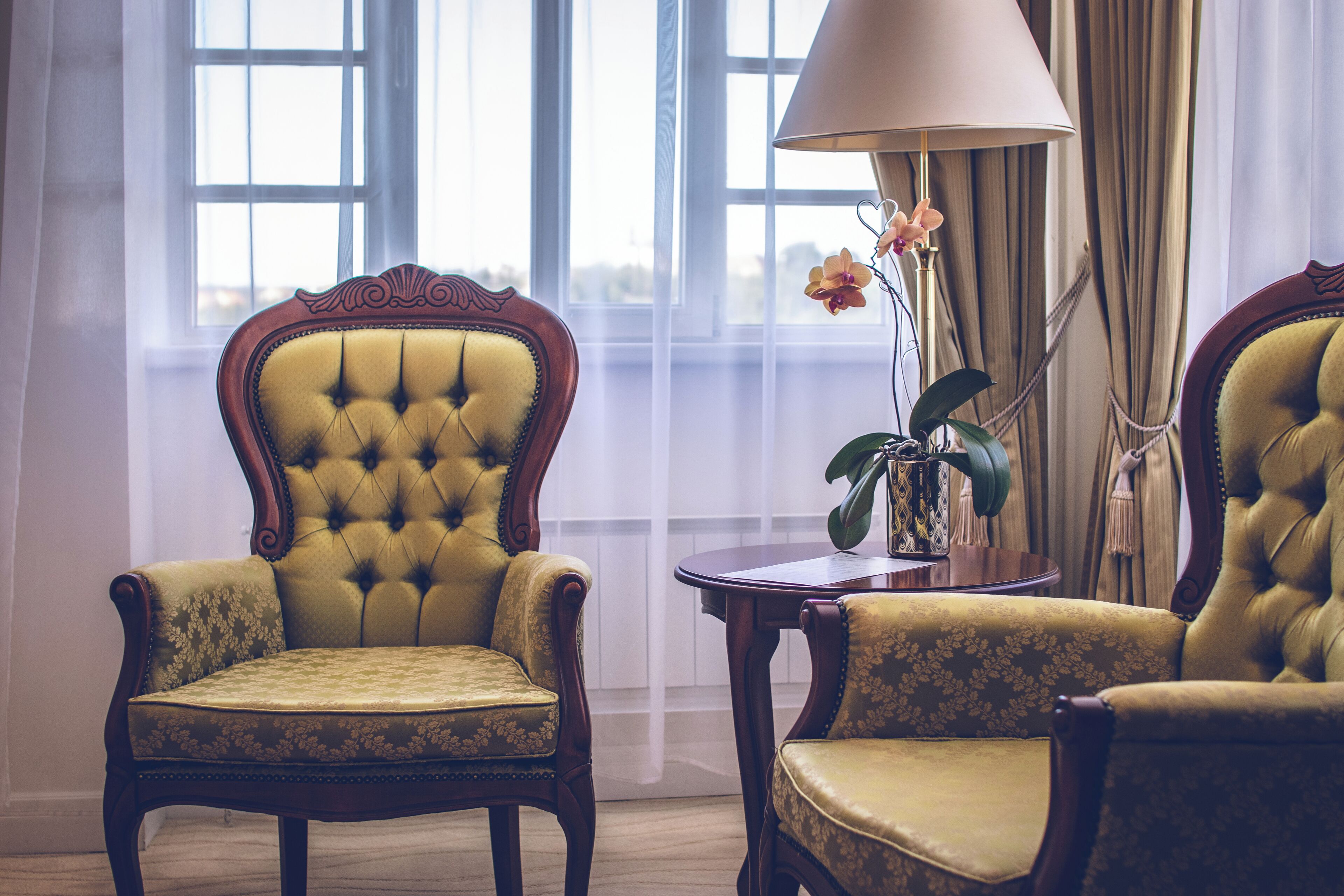 A room with two ornate chairs and a table, a lamp, and a vase with flowers.