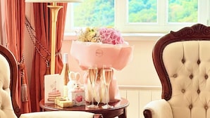Una habitación con vistas a un paisaje verde a través de una ventana, una mesa con una lámpara, una silla y un ramo de flores.
