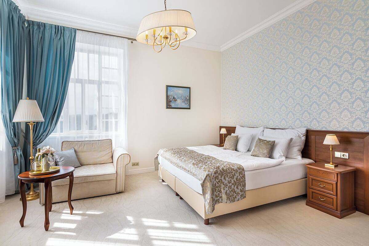A hotel room with a bed, a sofa, a nightstand, a lamp, a picture on the wall, and a window with curtains.