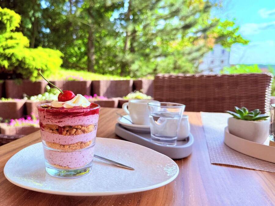 Un dessert composé de plusieurs couches, présenté sur une assiette en verre, accompagné d’une tasse et d’une soucoupe, d’un verre d’eau et d’une petite plante en pot, le tout sur une table à l’extérieur.