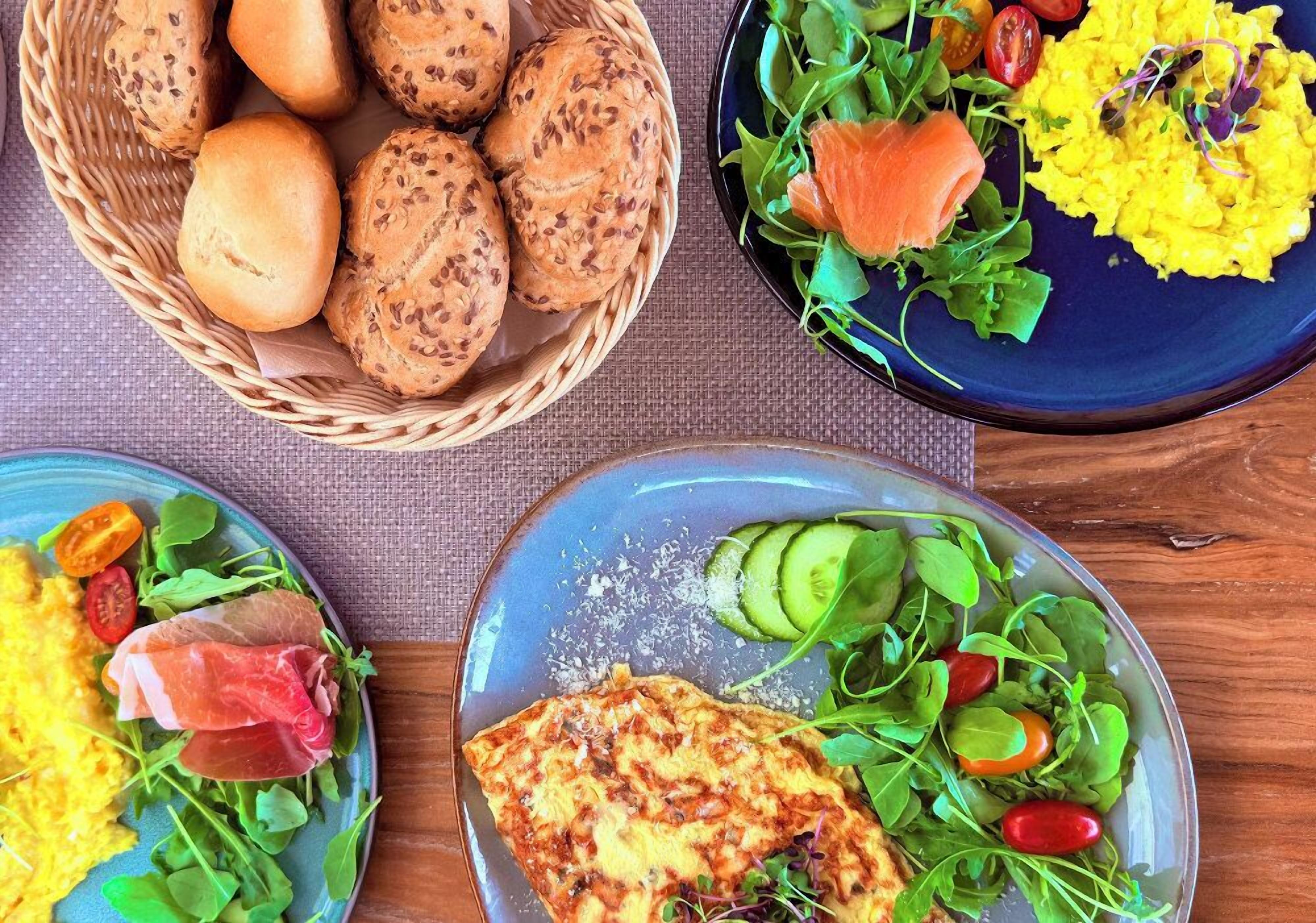 Una mesa con tres platos de comida, incluyendo bollos de pan, una ensalada con salmón y huevo, y un plato con pechuga de pollo a la parrilla y vegetales verdes.