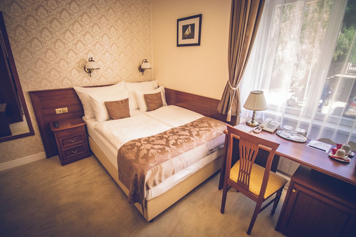 A hotel room with a bed, a desk with a chair, a lamp, and a window with curtains.