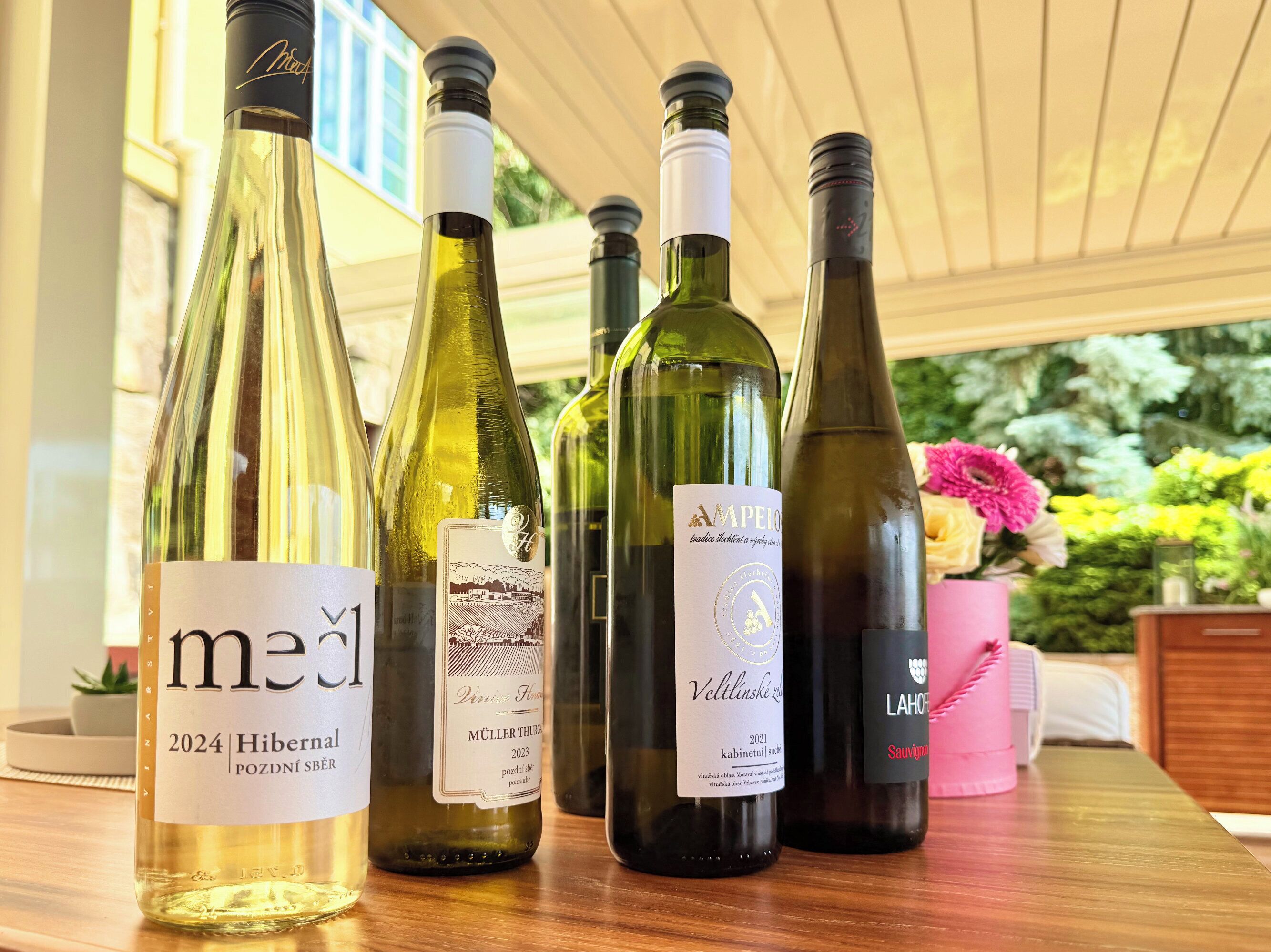 A selection of wine bottles on a wooden table with a view of greenery and a pink vase in the background.