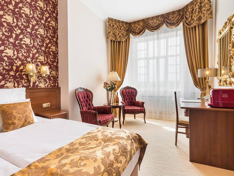 Una habitación de hotel con una cama, dos sillones rojos, un escritorio, una lámpara y un espejo.