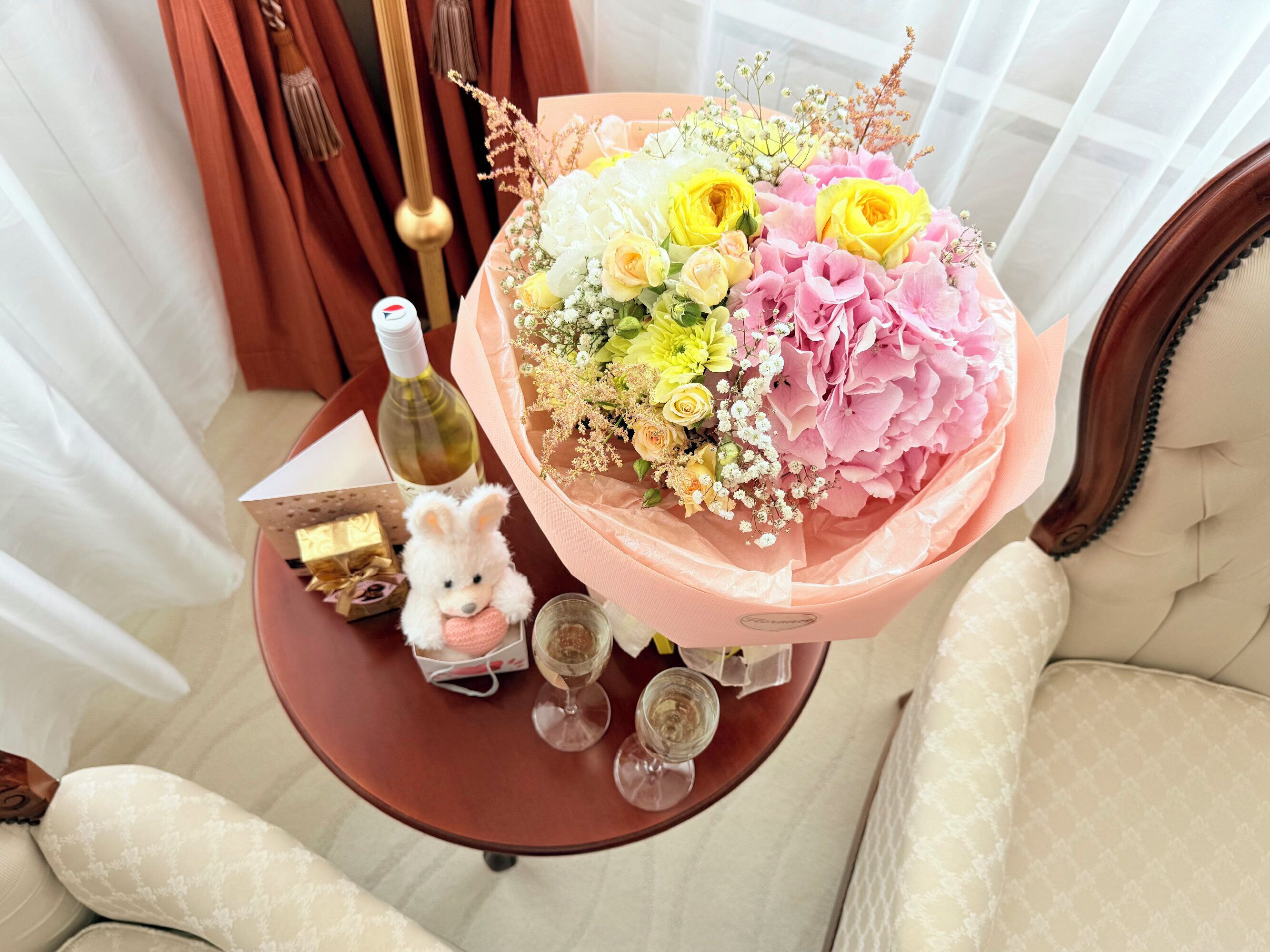 Una mesa con un ramo de flores, una botella de vino, un osito de peluche y dos copas.