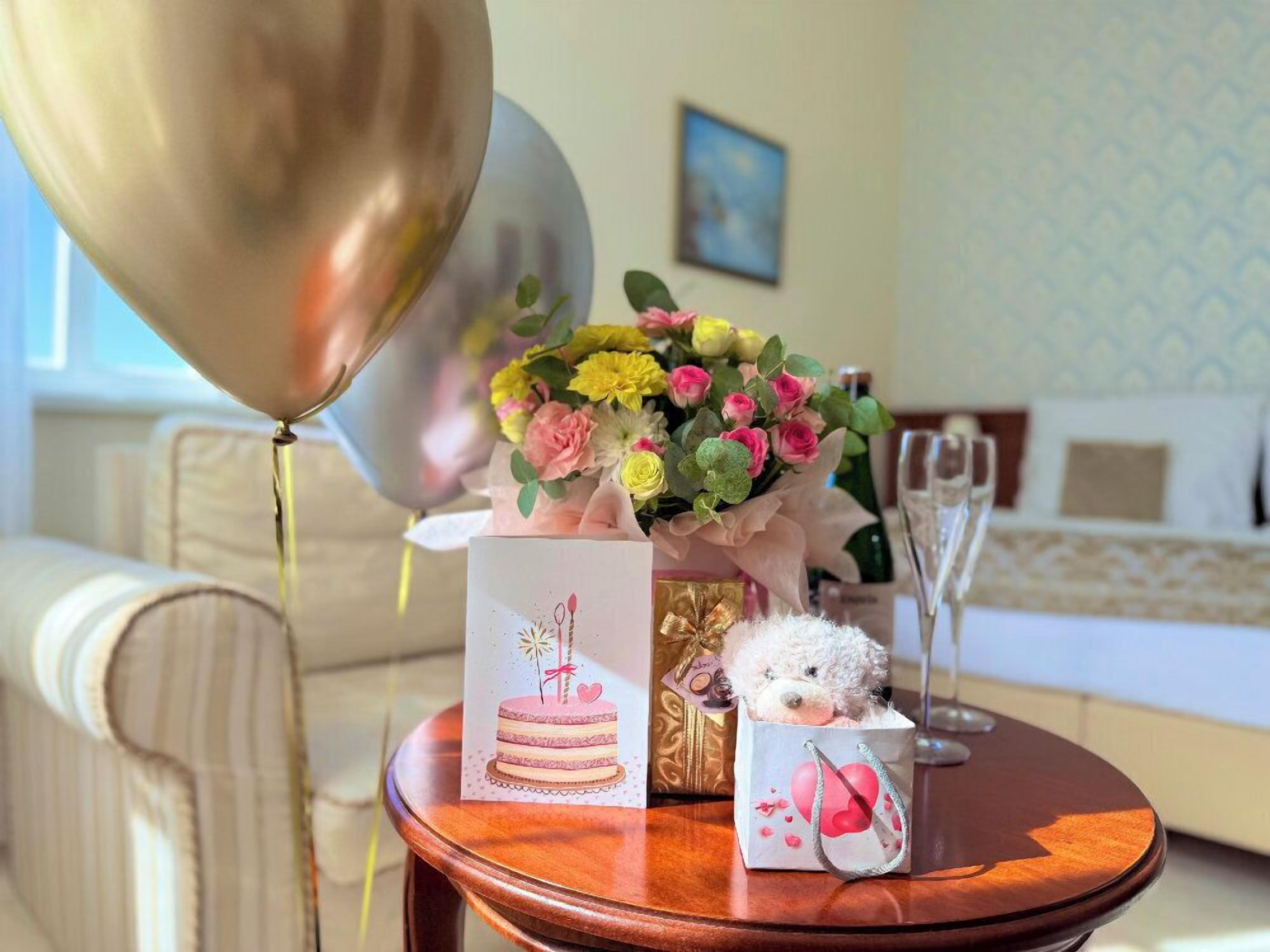 A table with a bouquet of flowers, a teddy bear, a cake illustration, a gift box, and a champagne glass.
