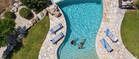 An aerial view of a swimming pool with a curved design, surrounded by stone tiles and equipped with lounge chairs.