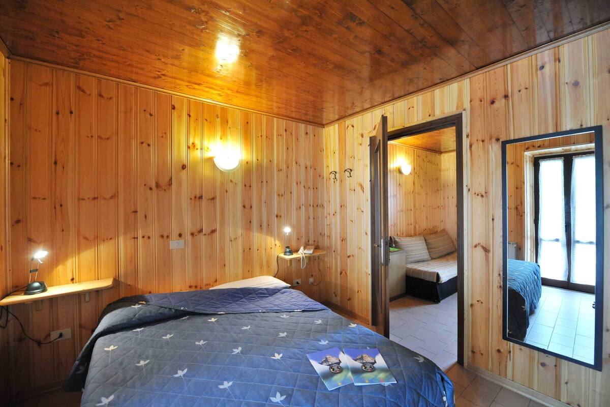 A wooden-paneled room with a bed, bedside table, and a mirror reflecting a window.