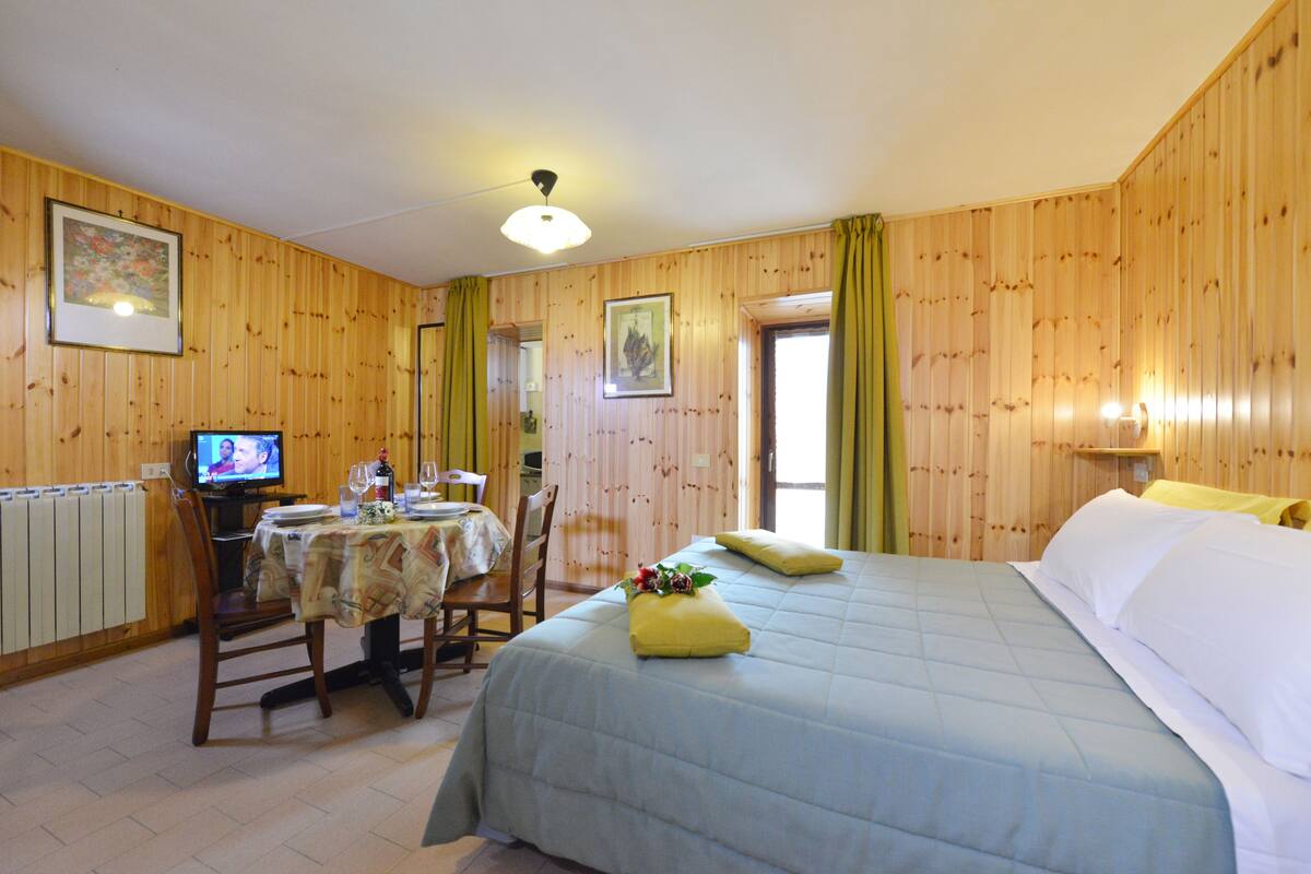A room with wooden paneling, a bed with a blue bedspread, a table with a tablecloth, a chair, a television, and a window with curtains.