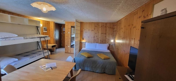 A room with bunk beds, a desk, a chair, a bed with pillows, a lamp, and a television.