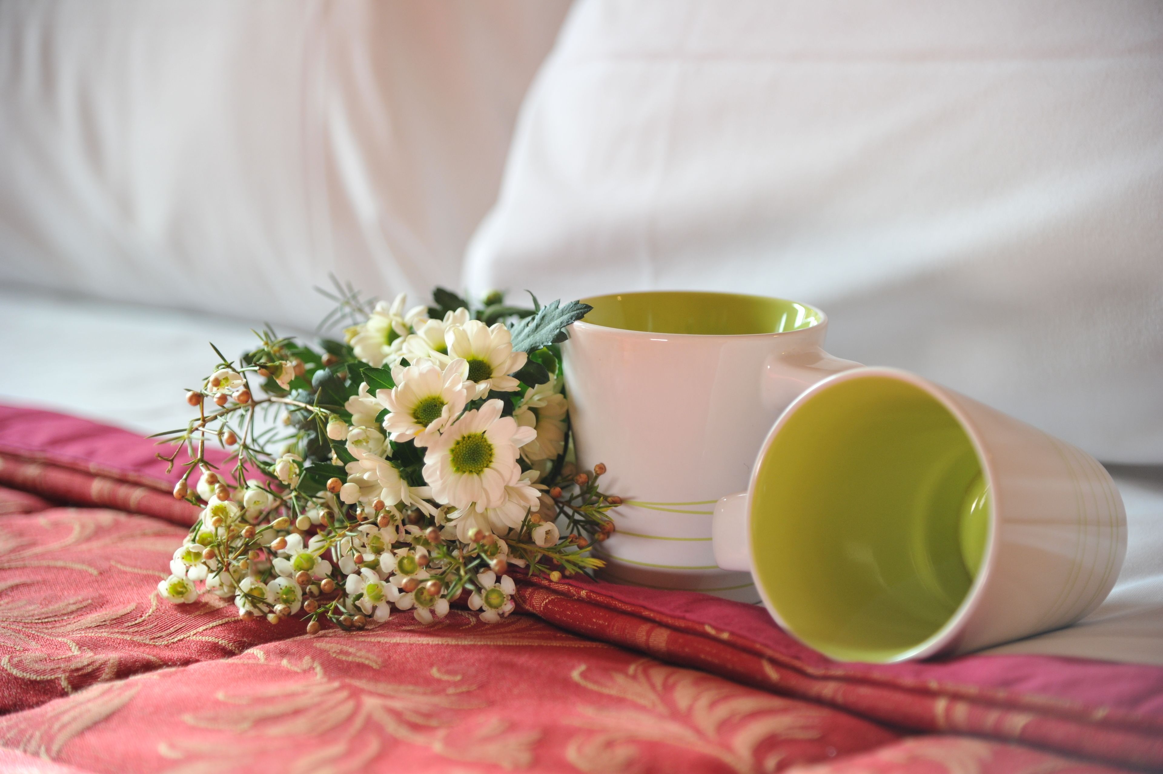 A bed with a white pillow, a green mug, and a bouquet of white flowers on a patterned red blanket.