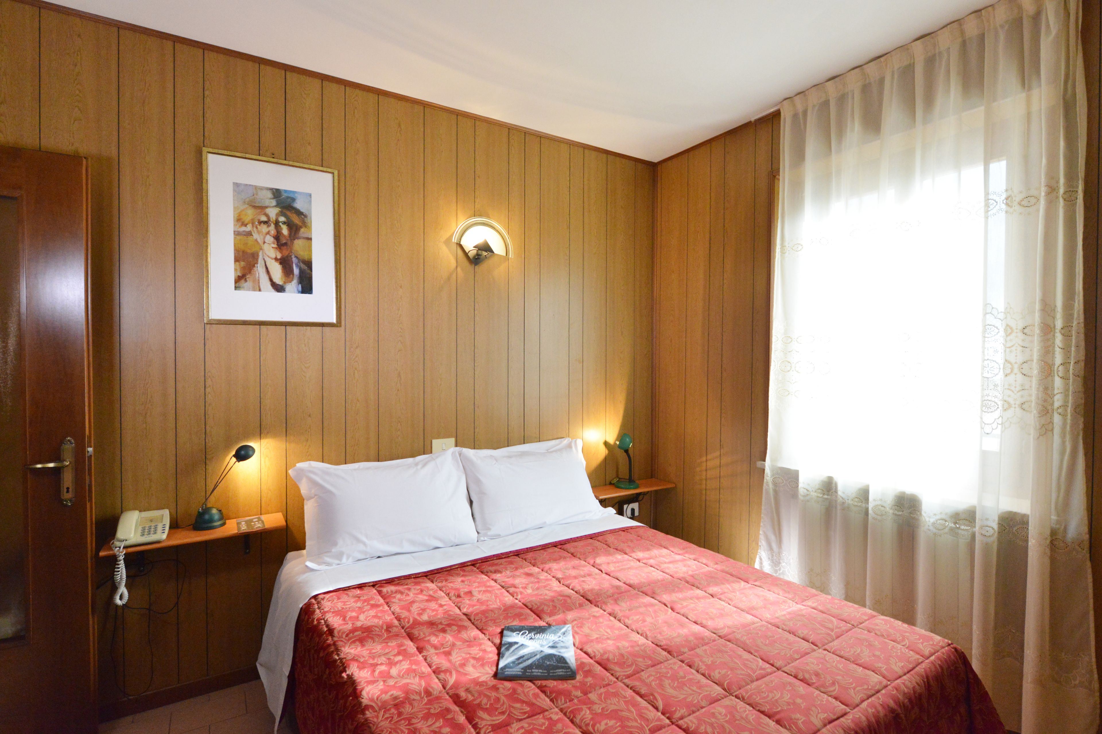 Une chambre d’hôtel avec un mur lambrissé en bois, un lit recouvert d’une courtepointe rouge, une lampe de chevet, un tableau encadré et une fenêtre ornée de rideaux légers.