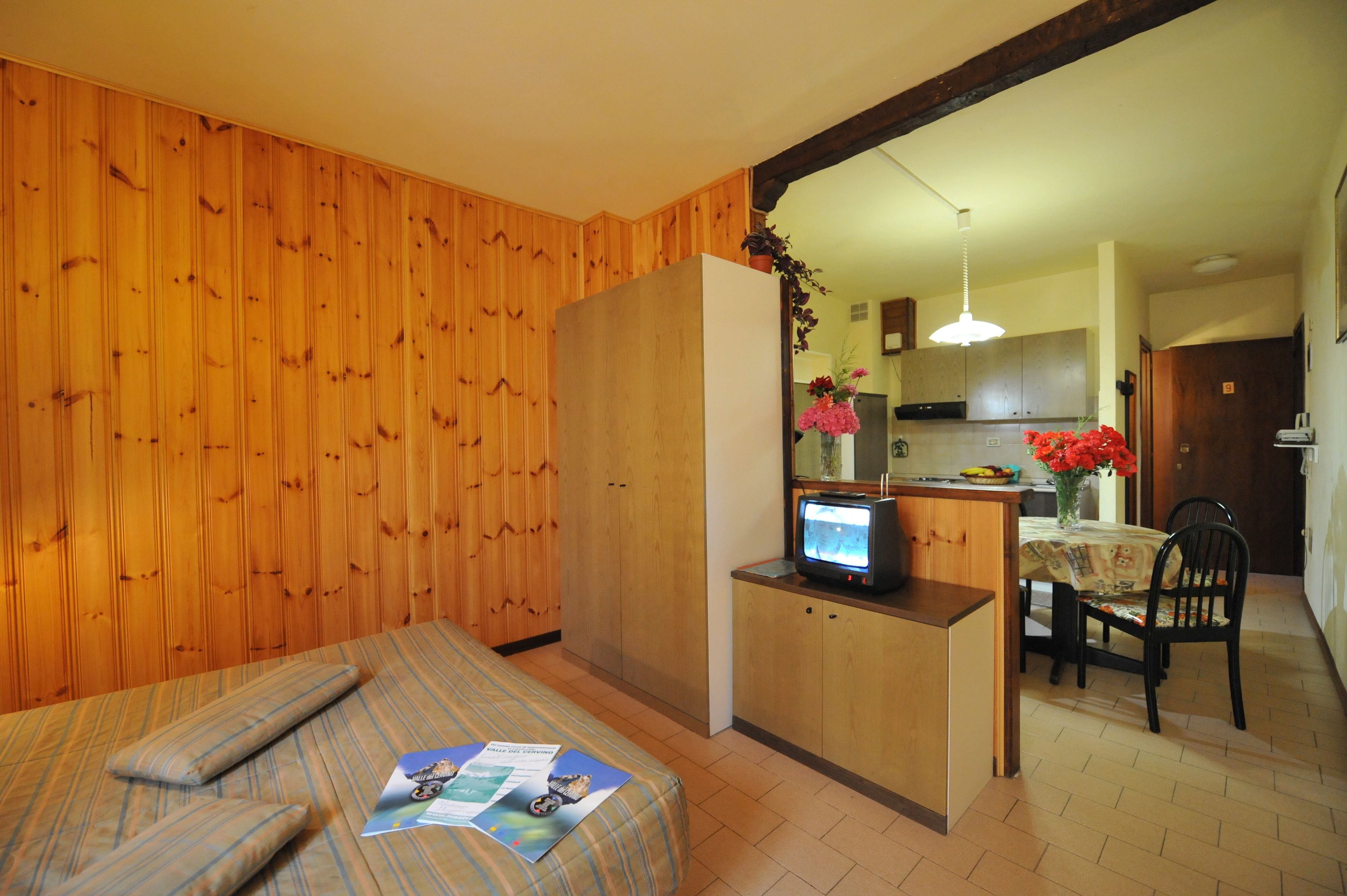 A room with wooden walls, a bed with a striped blanket, a small kitchenette with a TV, and a dining area with a table and chairs.