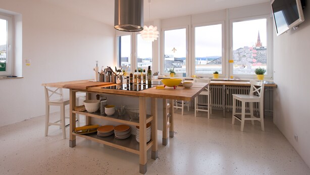 Une cuisine moderne dotée d’un îlot central, de chaises blanches et de grandes fenêtres offrant une vue sur un pont et le paysage urbain.