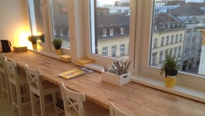Une table en bois, des chaises blanches, une fenêtre donnant sur des immeubles et une plante posée sur le rebord de la fenêtre.