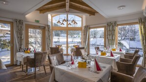 Un restaurant avec de nombreuses tables dressées pour le service, de grandes fenêtres donnant sur un paysage enneigé et un plafond en bois.