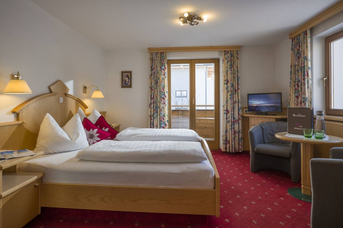 A hotel room with two beds, a TV, a small table with glasses, and a view of the outside through a door.