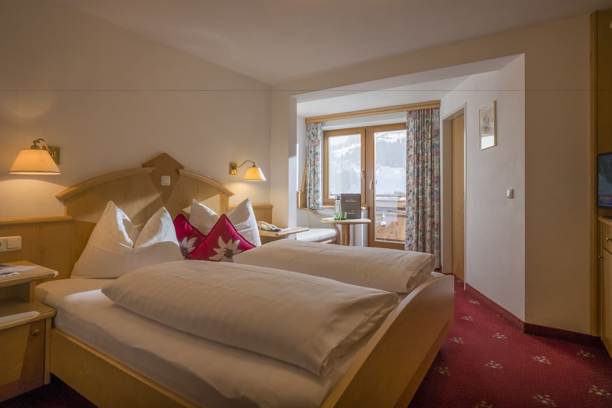 A hotel room with a large bed, two bedside tables, a small table with a lamp, a television, and a window with curtains.
