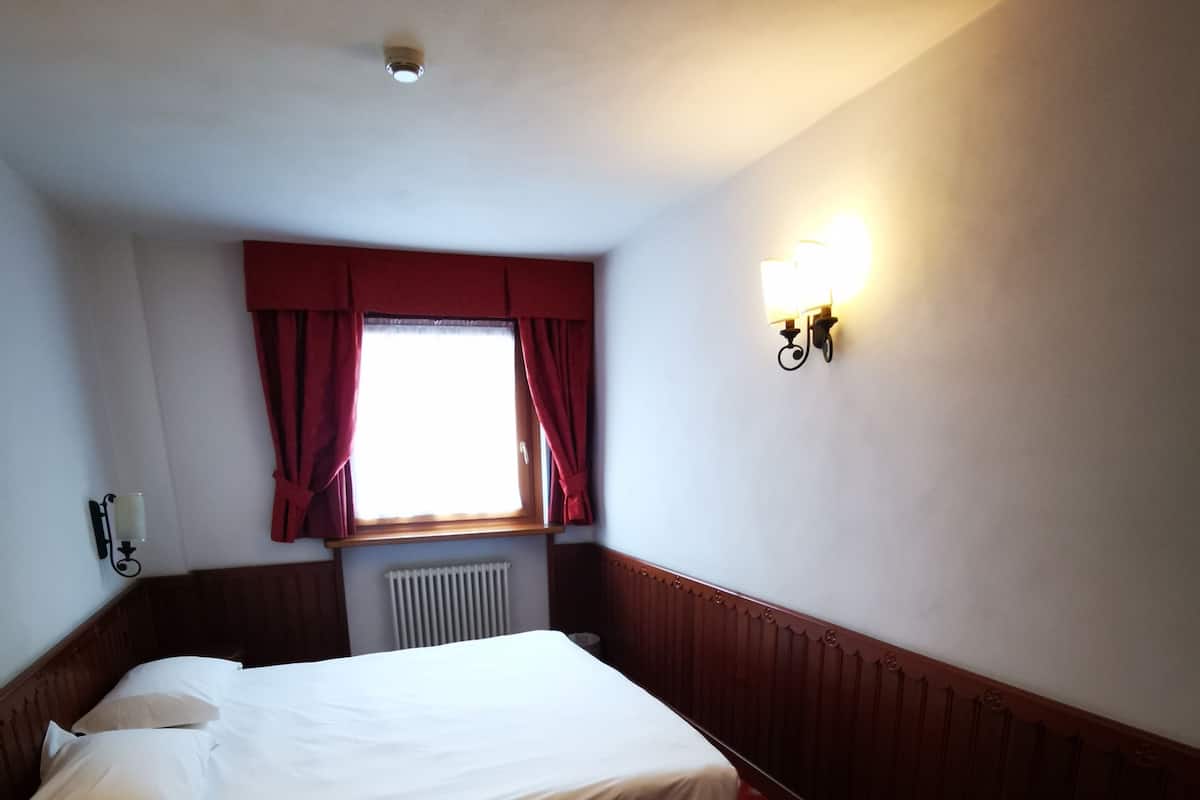 Ein Hotelzimmer mit einem Einzelbett, einem Nachttisch aus Holz, einem Schreibtisch, einem Fenster mit roten Vorhängen und einer wandmontierten Lampe.