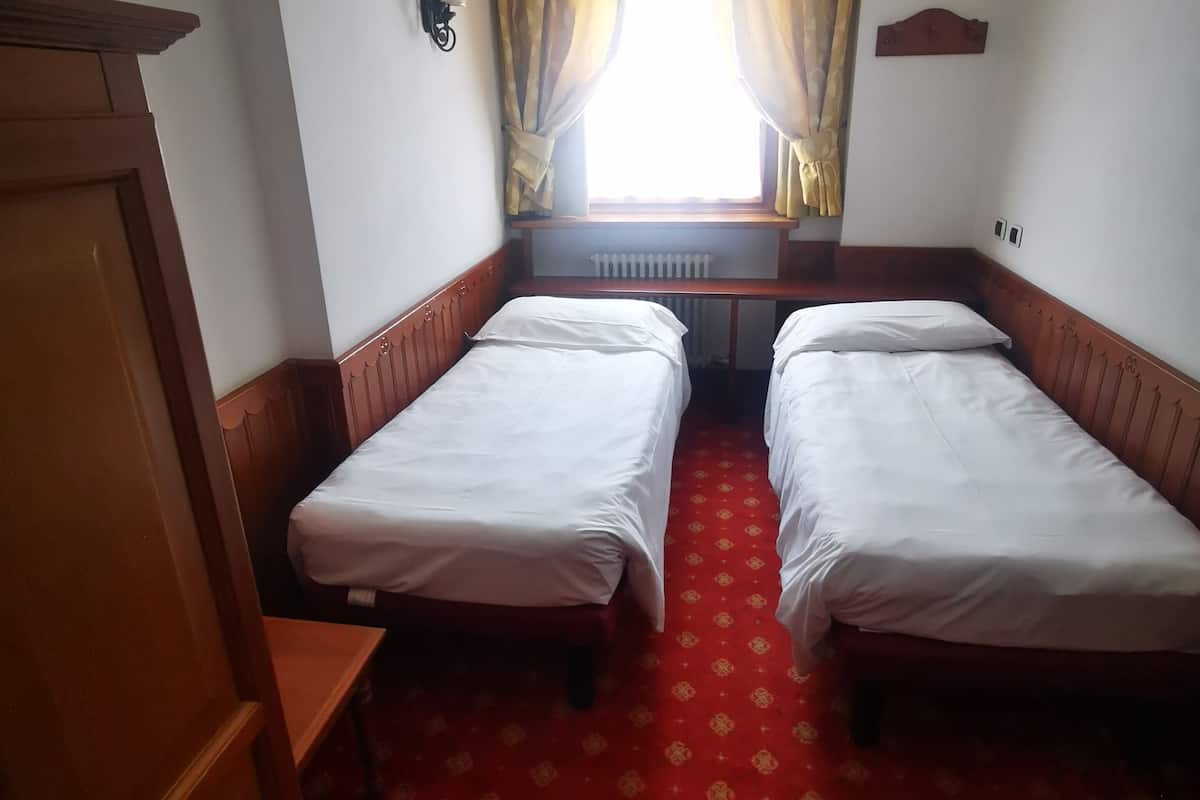 Ein Hotelzimmer mit zwei Einzelbetten, einem Holzschrank, einem Heizkörper und einem mit Vorhängen versehenen Fenster.