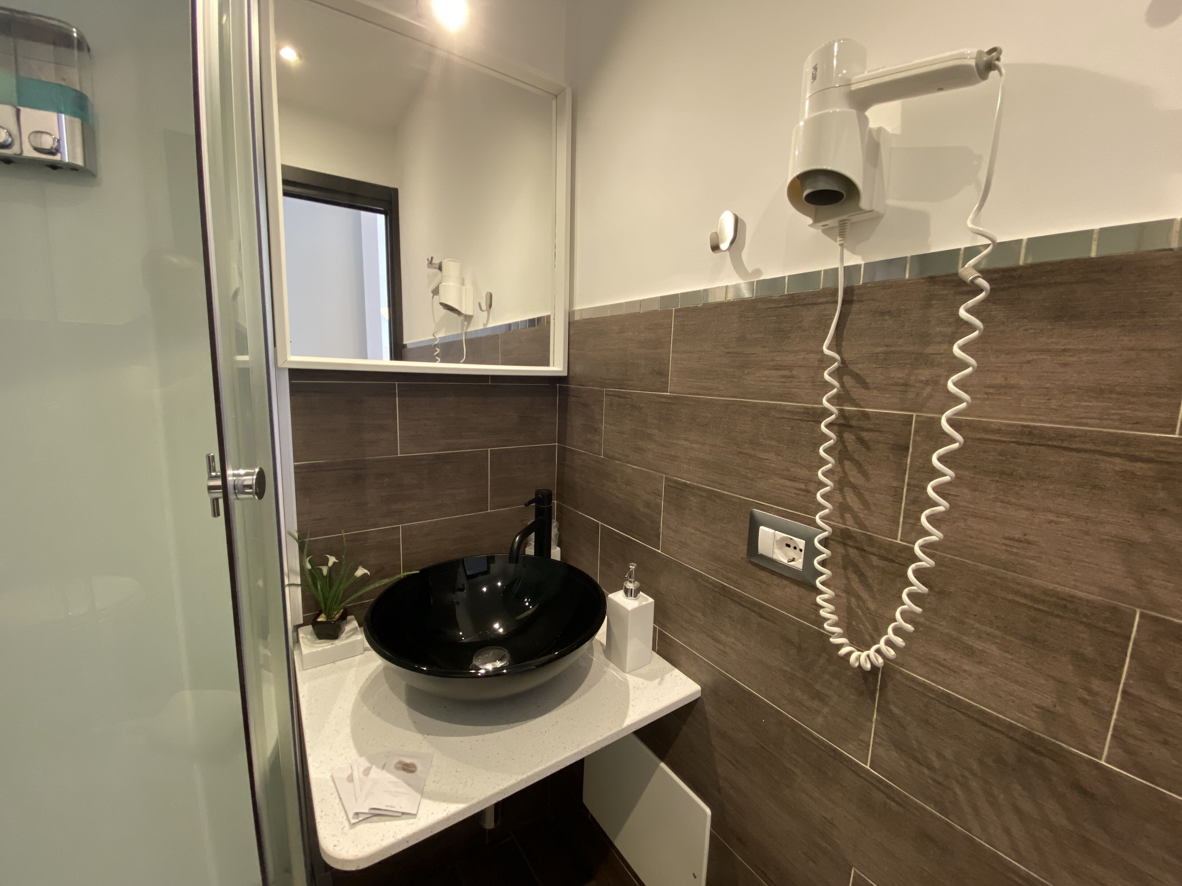 Un baño moderno con cabina de ducha de vidrio, un lavamanos negro y un secador de pelo fijado a la pared.