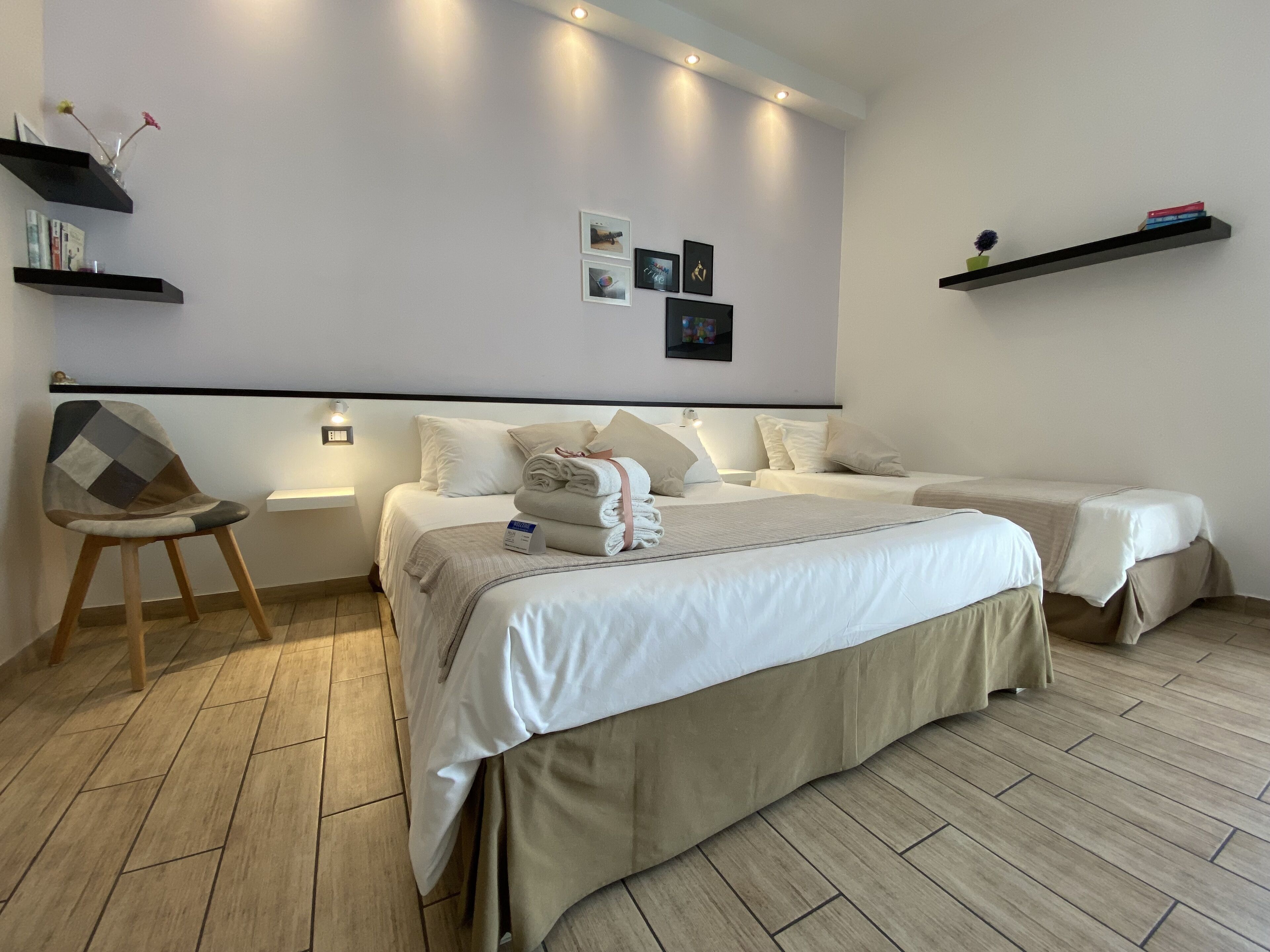 A hotel room with two beds, a chair, and shelves with books and decorative items.
