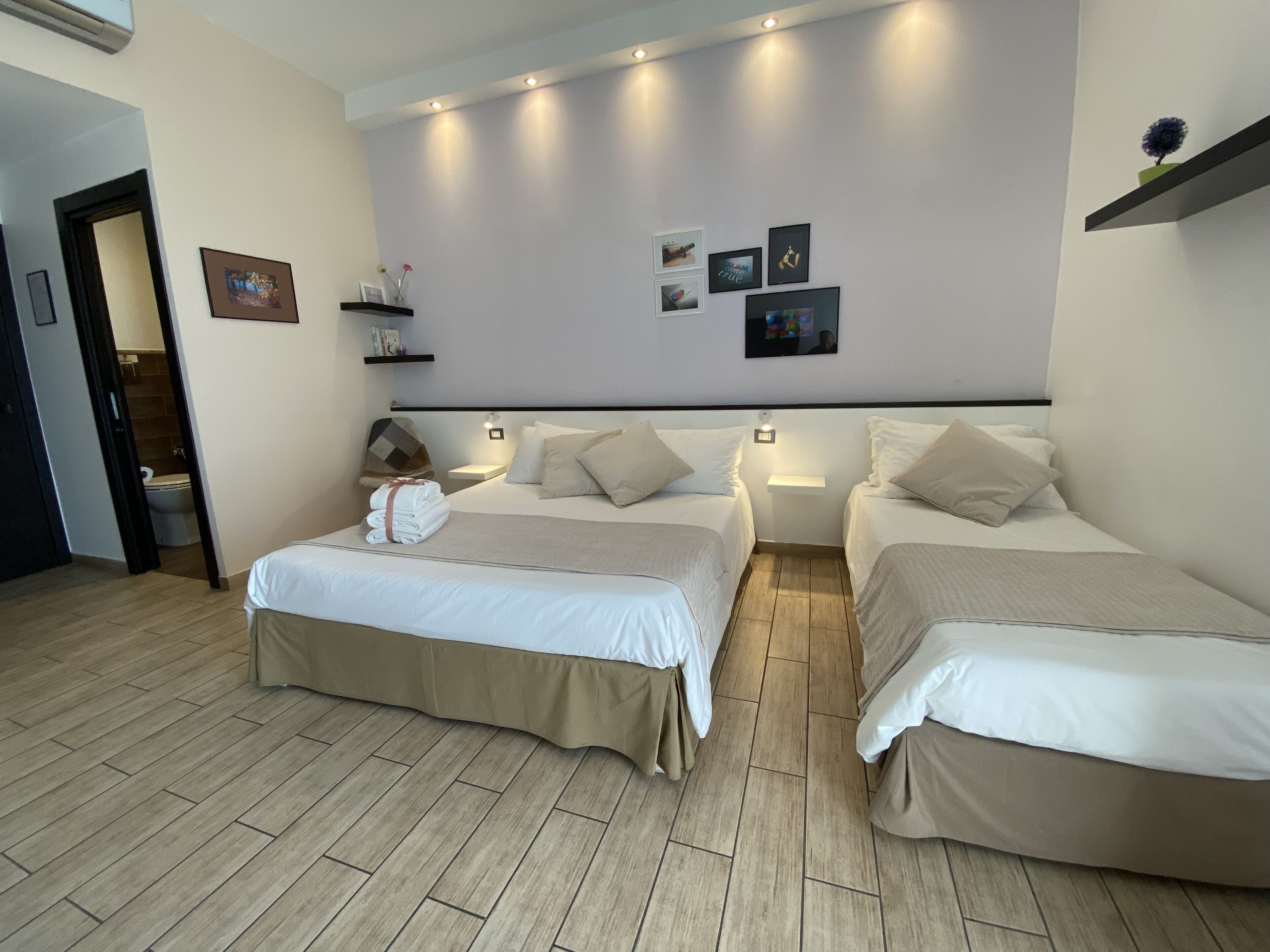 A hotel room with two beds, a wooden floor, a wall-mounted shelf with books and a plant, and a door leading to a bathroom.