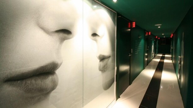 Ein Korridor mit schwarz-weiß gestreiftem Boden, grünen Wänden und einer großen Spiegelwand, die teilweise ein Gesicht reflektiert.